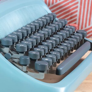 Puede incluir: Una m&aacute;quina de escribir vintage azul claro con teclas grises. Las teclas est&aacute;n dispuestas en un dise&ntilde;o QWERTY est&aacute;ndar. La m&aacute;quina de escribir tiene una barra espaciadora gris y una tecla roja. El fondo es un patr&oacute;n de rayas rojas y blancas.