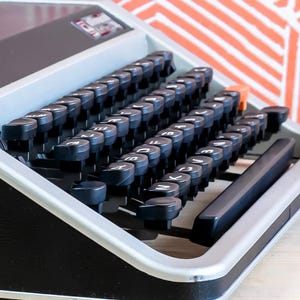 Puede incluir: Primer plano de una m&aacute;quina de escribir vintage con teclas negras y un marco plateado. Las teclas est&aacute;n dispuestas en un dise&ntilde;o QWERTY est&aacute;ndar. La m&aacute;quina de escribir tiene un cuerpo negro y est&aacute; sobre una superficie clara.