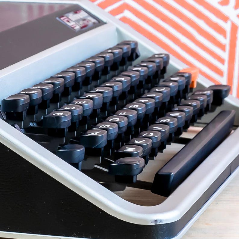 Puede incluir: Primer plano de una m&aacute;quina de escribir vintage con teclas negras y un marco plateado. Las teclas est&aacute;n dispuestas en un dise&ntilde;o QWERTY est&aacute;ndar. La m&aacute;quina de escribir tiene un cuerpo negro y est&aacute; sobre una superficie clara.