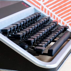 Puede incluir: Primer plano de una m&aacute;quina de escribir vintage con teclas negras y un marco plateado. Las teclas est&aacute;n dispuestas en un dise&ntilde;o QWERTY est&aacute;ndar. La m&aacute;quina de escribir est&aacute; sobre una superficie de madera, con un fondo naranja y blanco estampado.