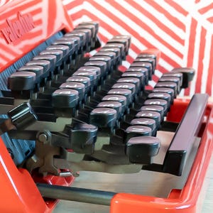 Puede incluir: Primer plano de una m&aacute;quina de escribir vintage roja con teclas negras. Las teclas est&aacute;n dispuestas en filas y tienen letras blancas. La m&aacute;quina de escribir tiene un acabado brillante y se encuentra sobre un fondo a rayas rojas y blancas. La palabra "Valentine" es visible.