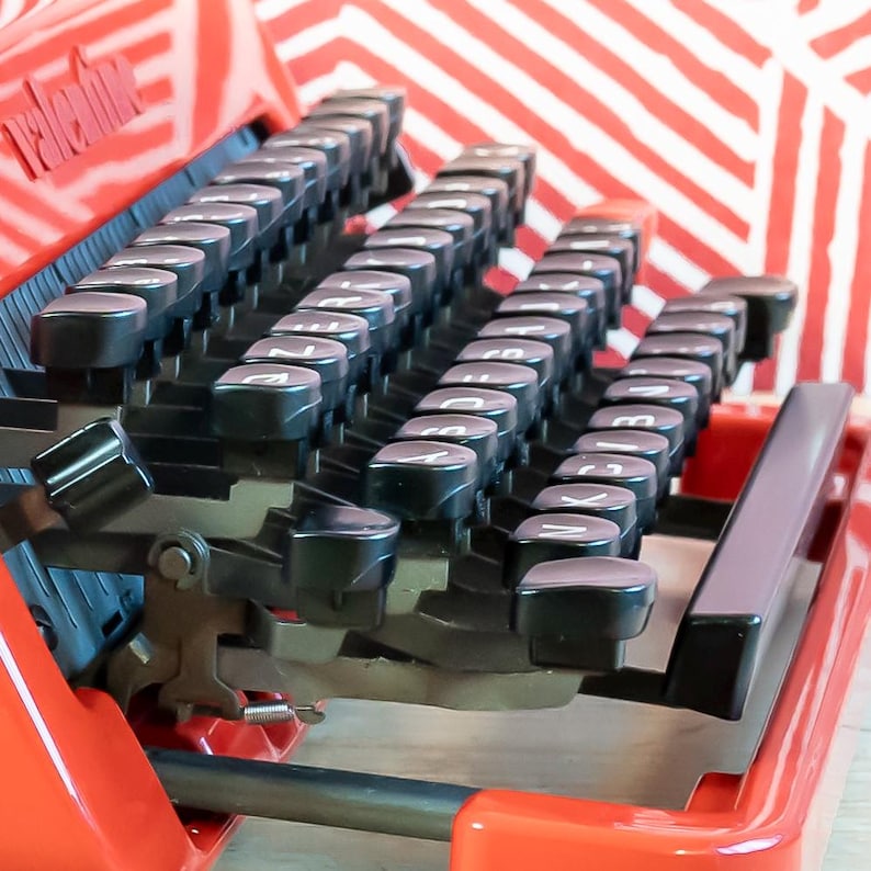 Puede incluir: Primer plano de una m&aacute;quina de escribir vintage roja con teclas negras. Las teclas est&aacute;n dispuestas en filas y tienen letras blancas. La m&aacute;quina de escribir tiene un acabado brillante y se encuentra sobre un fondo a rayas rojas y blancas. La palabra "Valentine" es visible.