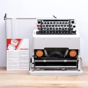 May include: A white Olivetti Valentine portable typewriter with a QWERTY keyboard, black circular keys, and a single red key. It features two orange spools, a metal carriage, and comes with a white carrying case and an instructional manual.