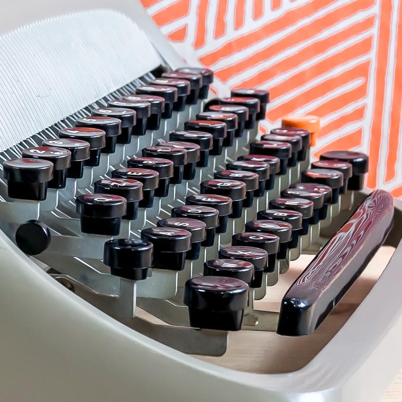 Puede incluir: Primer plano de una m&aacute;quina de escribir vintage con teclas negras y cuerpo gris. Las teclas est&aacute;n dispuestas en filas, con letras y n&uacute;meros visibles. La barra espaciadora y el retorno de carro tambi&eacute;n son visibles. El fondo presenta un dise&ntilde;o de patr&oacute;n naranja y blanco.