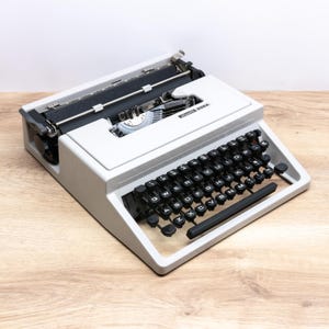 Pode incluir: Uma máquina de escrever Olivetti Dora cinza-claro com teclado QWERTY, teclas circulares pretas, carro de metal e fita preta. O nome da marca Olivetti Dora está impresso em preto no lado superior direito da carcaça.