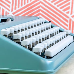 Puede incluir: Primer plano de una m&aacute;quina de escribir turquesa vintage con teclas blancas. Las teclas est&aacute;n dispuestas en filas y la barra espaciadora es visible. El fondo presenta un patr&oacute;n geom&eacute;trico coral y blanco.