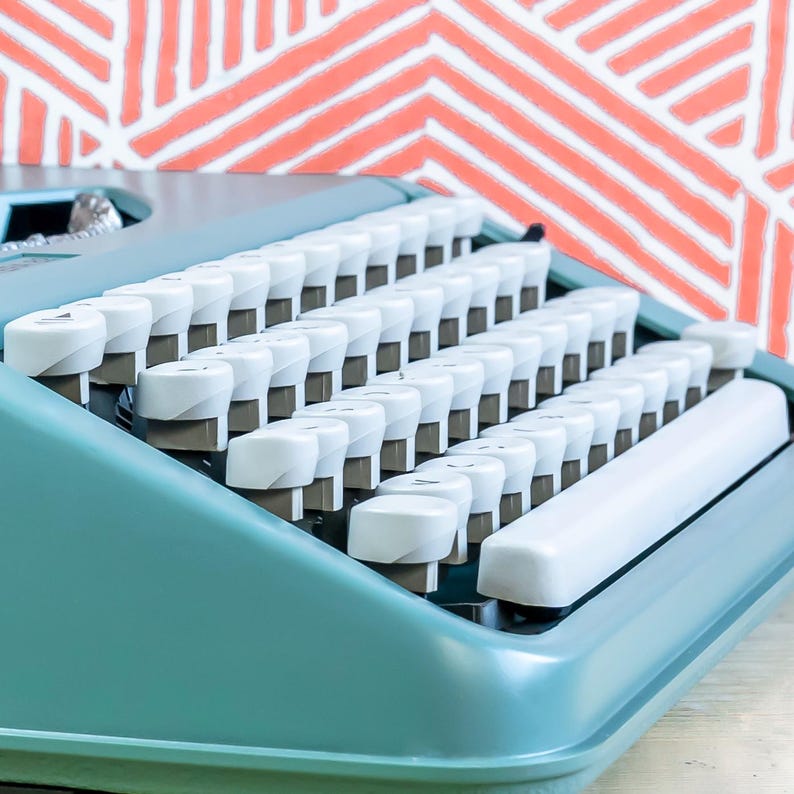 Puede incluir: Primer plano de una m&aacute;quina de escribir turquesa vintage con teclas blancas. Las teclas est&aacute;n dispuestas en filas y la barra espaciadora es visible. El fondo presenta un patr&oacute;n geom&eacute;trico coral y blanco.