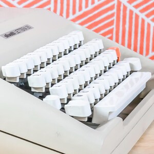 Puede incluir: Primer plano de una m&aacute;quina de escribir Olivetti Lettera 35. La m&aacute;quina de escribir tiene teclas blancas y un cuerpo gris claro. Las teclas est&aacute;n dispuestas en filas, con algunas teclas naranjas. El fondo es un patr&oacute;n geom&eacute;trico en naranja y blanco.