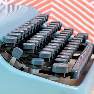 Puede incluir: Primer plano de una m&aacute;quina de escribir azul vintage con teclas grises. Las teclas est&aacute;n dispuestas en filas y la palanca de retorno del carro es visible. El fondo presenta un patr&oacute;n en zigzag rojo y blanco.