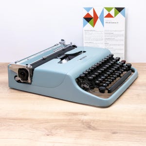 May include: A light blue Olivetti Lettera 22 portable typewriter with a black keyboard and metal carriage components. The typewriter is positioned in front of a white informational card featuring a geometric pattern and the text Olivetti Lettera 22.