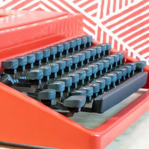 Puede incluir: Primer plano de una m&aacute;quina de escribir roja con un teclado completo de teclas grises. Las teclas est&aacute;n dispuestas en filas, con n&uacute;meros y letras visibles. La m&aacute;quina de escribir est&aacute; sobre un fondo con estampado rojo y blanco.