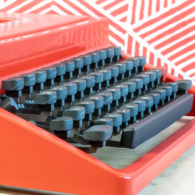 Puede incluir: Primer plano de una m&aacute;quina de escribir roja con un teclado completo de teclas grises. Las teclas est&aacute;n dispuestas en filas, con n&uacute;meros y letras visibles. La m&aacute;quina de escribir est&aacute; sobre un fondo con estampado rojo y blanco.