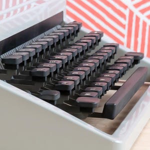 Puede incluir: Primer plano de una m&aacute;quina de escribir vintage con teclas negras y un cuerpo gris. Las teclas est&aacute;n dispuestas en un dise&ntilde;o curvo, con letras y s&iacute;mbolos visibles. La barra espaciadora es de color marr&oacute;n oscuro. El fondo presenta un patr&oacute;n geom&eacute;trico rojo y blanco.