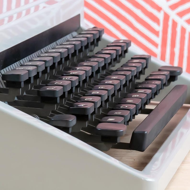 Puede incluir: Primer plano de una m&aacute;quina de escribir vintage con teclas negras y un cuerpo gris. Las teclas est&aacute;n dispuestas en un dise&ntilde;o curvo, con letras y s&iacute;mbolos visibles. La barra espaciadora es de color marr&oacute;n oscuro. El fondo presenta un patr&oacute;n geom&eacute;trico rojo y blanco.