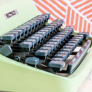 Puede incluir: Una m&aacute;quina de escribir verde menta vintage con teclas grises. Las teclas est&aacute;n dispuestas en un dise&ntilde;o QWERTY est&aacute;ndar. La m&aacute;quina de escribir tiene una etiqueta que dice "Lettera 32". El fondo presenta un dise&ntilde;o blanco y naranja.