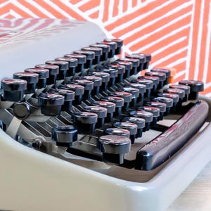 Puede incluir: Primer plano de una m&aacute;quina de escribir vintage con teclas negras y un cuerpo gris claro. Las teclas est&aacute;n dispuestas en filas, con las letras y los n&uacute;meros visibles en cada tecla. El fondo presenta un patr&oacute;n geom&eacute;trico naranja y blanco.