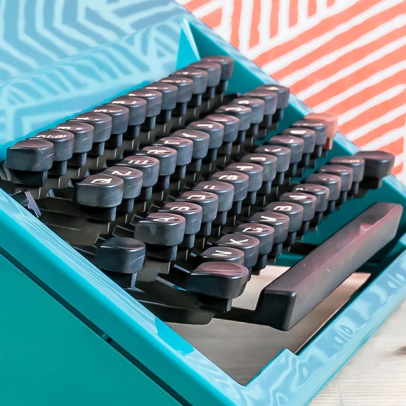 Puede incluir: Una m&aacute;quina de escribir vintage turquesa con teclas negras y una carcasa turquesa a juego. Las teclas est&aacute;n dispuestas en un dise&ntilde;o QWERTY est&aacute;ndar. El fondo presenta un patr&oacute;n de rayas blancas y naranjas.
