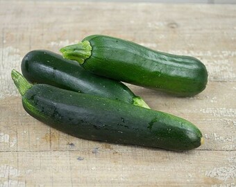 May include: Three dark green zucchini squash on a light wood surface.