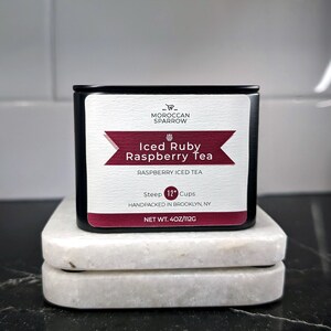 May include: A black tin container of Iced Ruby Raspberry Tea, a raspberry iced tea blend from Moroccan Sparrow. The label is red and white with the text "Iced Ruby Raspberry Tea", "Raspberry Iced Tea", "Steep 12 Cups", "Handpacked in Brooklyn, NY", and "Net Wt. 4oz/112g".
