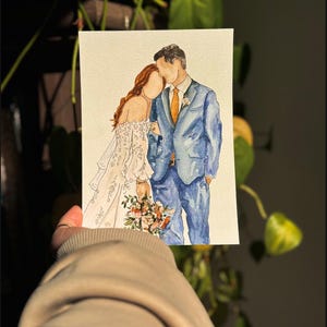 May include: Watercolor painting of a couple in wedding attire. The bride is wearing a white dress with lace detailing and the groom is wearing a blue suit with a light orange tie. They are standing close together and the bride is holding a bouquet of flowers.