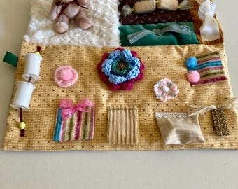 May include: A sensory activity mat with various textures and elements. Features a small teddy bear, a fluffy white square, and a yellow gingham patterned fabric. Includes a blue and pink crocheted flower, a pink bow, and other tactile elements.