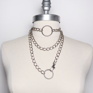 May include: A silver chain necklace with three layers and two large circular pendants. The necklace is displayed on a white mannequin.