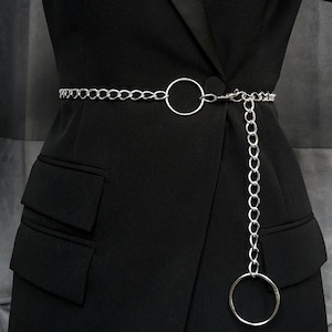 Un modèle vêtu de noir porte une ceinture en chaine argentée à la taille. Il y a deux anneaux ; un au centre de la ceinture et un autre, plus grand, pend perpendiculairement au bout de la chaine.