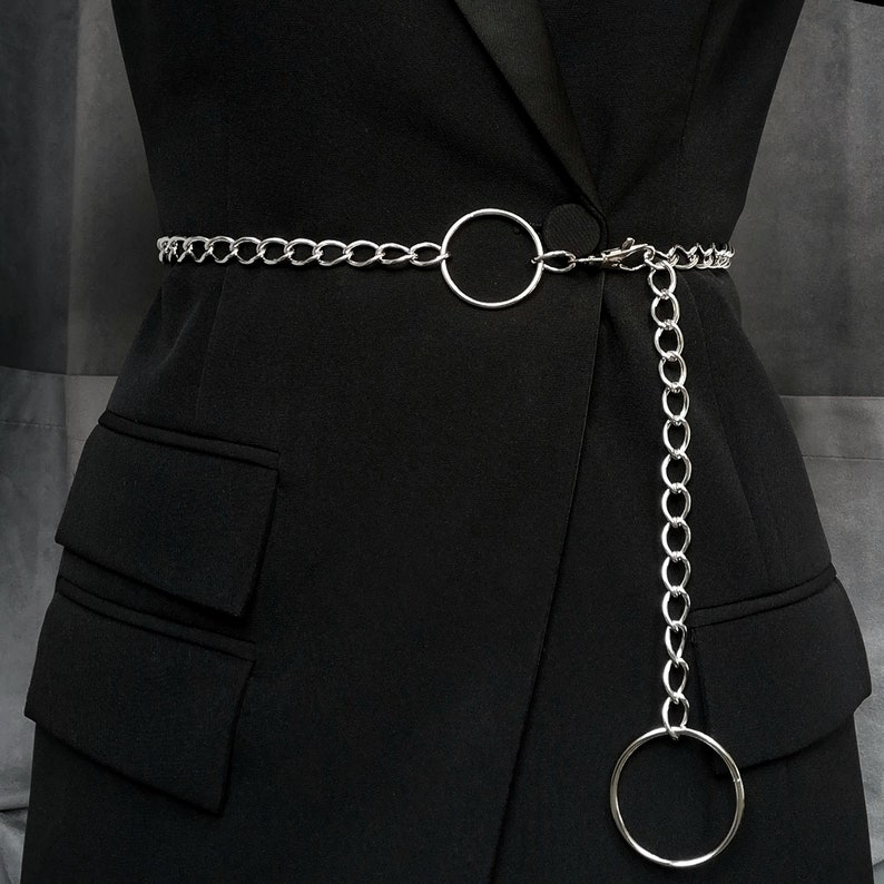 Un modèle vêtu de noir porte une ceinture en chaine argentée à la taille. Il y a deux anneaux ; un au centre de la ceinture et un autre, plus grand, pend perpendiculairement au bout de la chaine.