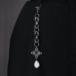 Op de afbeelding: Een zilverkleurig kettingaccessoire met een decoratieve kruisvormige hanger en een parelmoer druppel. De ketting is bevestigd aan een zwarte riem, waarschijnlijk voor een riem of tas. Het ontwerp is ingewikkeld en gedetailleerd.