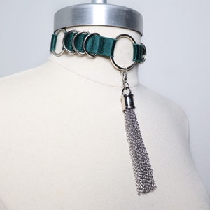 Puede incluir: Un collar gargantilla de terciopelo verde oscuro con múltiples anillos plateados y un borla de cadena plateada.