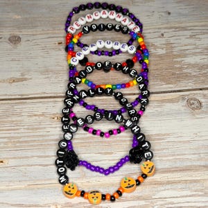 Puede incluir: Una colección de pulseras de cuentas con varias frases escritas con cuentas de letras blancas. Las pulseras presentan una mezcla de colores, incluyendo negro, morado, naranja y cuentas de arcoíris. Algunas pulseras tienen acentos de calabaza y rosa.
