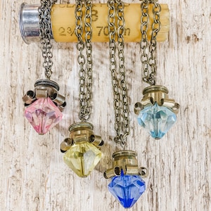 Puede incluir: Cuatro collares con colgantes de cristal de diferentes colores. Cada colgante está unido a una cadena de latón y a una caja de bala de latón. Los collares cuelgan de una superficie de madera.