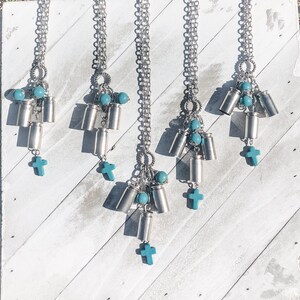 May include: Five silver-toned bullet casing pendants with turquoise beads and crosses. Each pendant hangs from a silver chain. The pendants are displayed against a white wooden background.