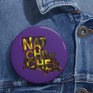 Peut inclure: Un bouton violet avec le texte "NATCHITOCHES" en or et vert, avec une carte de la Louisiane. Le bouton est posé sur une veste en jean bleue.
