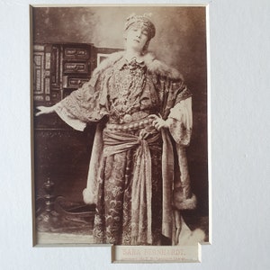May include: Sepia-toned photograph of a person in elaborate costume. The person wears a jeweled headdress, layered necklaces, and a patterned robe with fur trim. The image includes the text "SARA BERNHARDT" at the bottom.