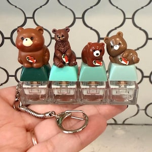 May include: A keychain featuring four mechanical keyboard switches topped with miniature brown bear figurines. Each bear holds a small red and white fish. The switches are mounted on a clear base, with two dark green and two light blue keycaps.