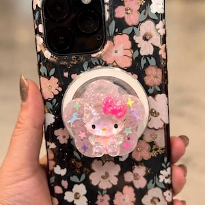 May include: A black smartphone case featuring a floral pattern with pink and white flowers and green leaves. Attached to the center is a circular phone grip with a translucent, glittery, flower-shaped base and a Hello Kitty figure with a bright pink bow.