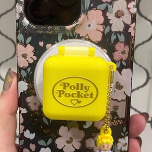 以下が含まれることがあります： 暗い背景にピンクと白の花柄が施されたスマートフォンケース。中央には明るい黄色の正方形のポーリーポケットのコンパクトアクセサリーが取り付けられており、金のチェーンに小さなフィギュアがぶら下がっています。