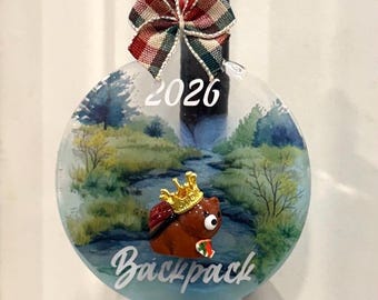 Peut inclure: Une décoration ronde et transparente ornée d'un paysage peint représentant un ruisseau et des arbres. Elle porte l'inscription 2026 et Backpack, avec une figurine d'ours brun portant une couronne dorée. Un nœud écossais est fixé au sommet.