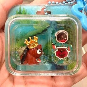 以下が含まれることがあります： 金色の王冠とバックパックを身に着けた茶色のクマのフィギュアが入った透明なプラスチック製キーホルダー。ケース内には小さなビーズと、赤い背景にクマのイラストとKatmai Conservancyの文字が書かれた円形のステッカーが入っています。