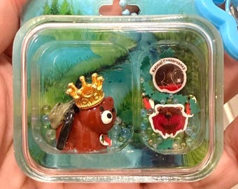 Può includere: Un portachiavi in plastica trasparente contenente una statuetta di orso bruno con una corona dorata, un piccolo zaino e un pesce. La custodia presenta uno sfondo forestale, piccole perline e due adesivi, uno dei quali reca Katmai Conservancy.