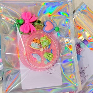 May include: A circular pink resin shaker charm filled with miniature figurines including a cake, a green dinosaur, and a sleeping character. The charm is attached to a beaded lanyard with a bright pink bow and is packaged in a holographic pouch.