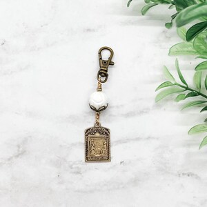 May include: A gold-toned keychain with a white bead and a rectangular charm featuring a religious image. The charm has the words "Santissima Trinita" inscribed on it.