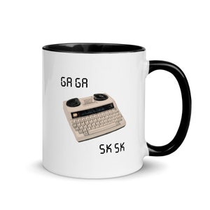 May include: White ceramic coffee mug with a black rim and handle. The mug features a black and white illustration of a vintage typewriter with the text "GA GA" above and "SK SK" below the image.