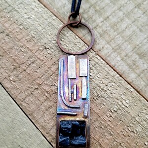 May include: Artisan-crafted pendant necklace featuring a rectangular, iridescent metal pendant with geometric silver accents and a dark blue cord. The pendant has a copper-colored frame and a section of dark blue stones.