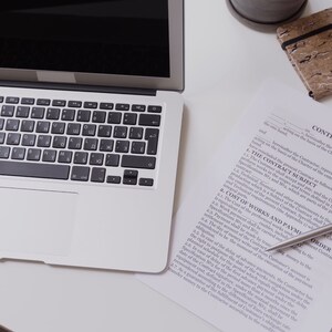 May include: A silver laptop with a black keyboard and a white trackpad is on a white surface. A contract with the text "CONTRACT NO" is next to a silver pen. A small notebook and a potted plant are also visible.