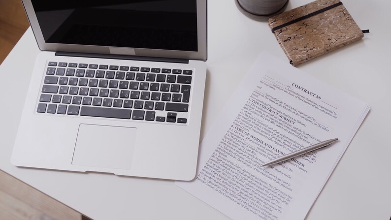 May include: A silver laptop with a black keyboard and a white trackpad is on a white surface. A contract with the text "CONTRACT NO" is next to a silver pen. A small notebook and a potted plant are also visible.
