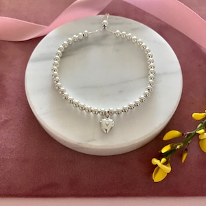 May include: A silver beaded bracelet with a small silver heart charm. The bracelet is on a white marble surface.