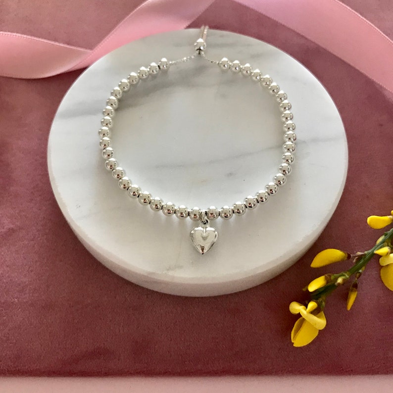 May include: A silver beaded bracelet with a small silver heart charm. The bracelet is on a white marble surface.