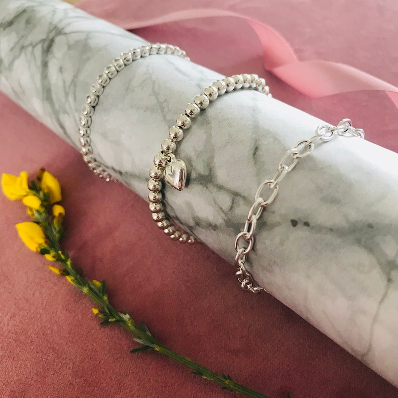 May include: Three silver bracelets on a marble surface. One bracelet is a chain link style, and the other two are beaded bracelets. One of the beaded bracelets has a small heart charm.